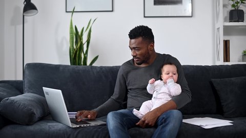 A young African-American father using laptop for remote work, while looking after his cute baby daughter. A freelancer dad with an infant