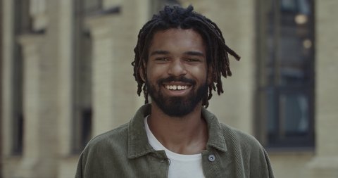 Crop view of handsome man with beard and moustache looking and posing to camera. Portrait of young guy with dreadlocks smiling while standing at street. Concept of lifestyle.