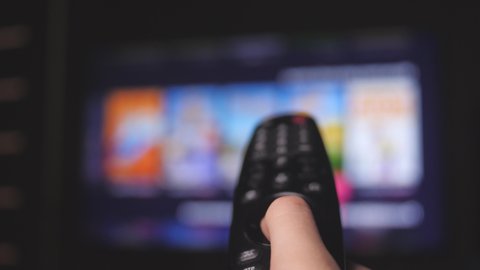 Man's hand selects internet tv channels with remote control, close-up. Person controls TV using a modern remote control. A man watches smart TV and uses black remote control. Blurry tv scrolls pages.