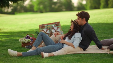 Young people chilling on blanket on green lawn at summer park. Smiling couple having rest on picnic on green meadow. Young man and woman laughing outdoors