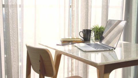 Beautiful workspace - online remote work from home concept. A wooden table with computer laptop, smartphone, coffee cup beside the window balcony with sun shine, wind through the curtain. No people.