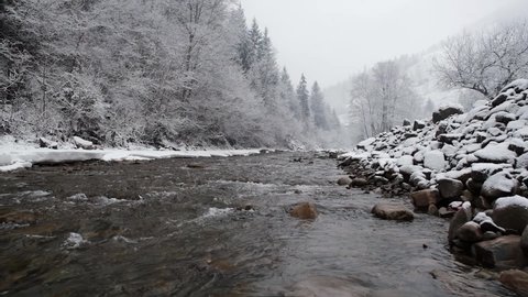 River flow with winter water runs at carpathian mountains, Ukraine. brook rounded with snow and trees at winter weather. Snow falling slow motion