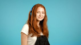 Pretty young girl with red hair listening to music, smiling, dancing in blue headphones in studio against plain background. Music, dance, radio concept, slow motion.