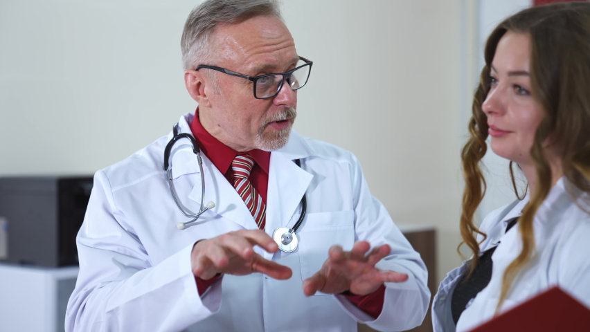 Serious focused middle aged physician in medical coat consults with woman doctor in clinic. Woman is listening to senior doctor at office. Royalty-Free Stock Footage #1065598168