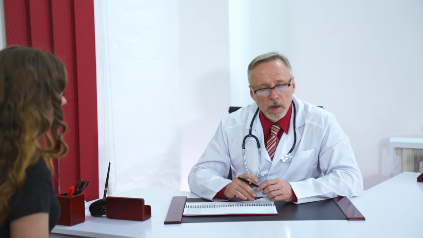 Side view of young female patient on consultation in clinic. Middle aged mature male doctor in white uniform. Health care concept. Royalty-Free Stock Footage #1065598174