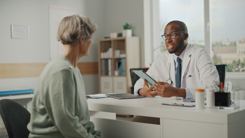 African American Family Doctor Showing Heart Analysis Results on Tablet Computer to Senior Female Patient During Consultation in a Health Clinic. Physician Sitting Behind a Desk in Hospital Office. | Shutterstock HD Video #1065599365