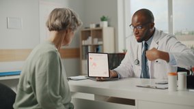 African American Family Doctor Showing Heart Analysis Results on Tablet Computer to Senior Female Patient During Consultation in a Health Clinic. Physician Sitting Behind a Desk in Hospital Office. - Powered by Shutterstock - Get 15% off with code: PIKWIZARD15