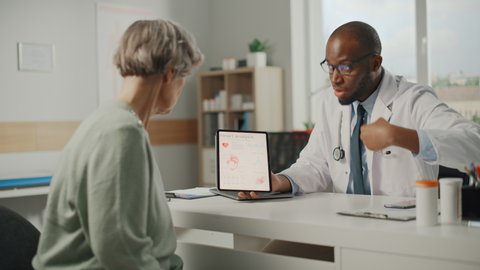 African American Family Doctor Showing Heart Analysis Results on Tablet Computer to Senior Female Patient During Consultation in a Health Clinic. Physician Sitting Behind a Desk in Hospital Office.