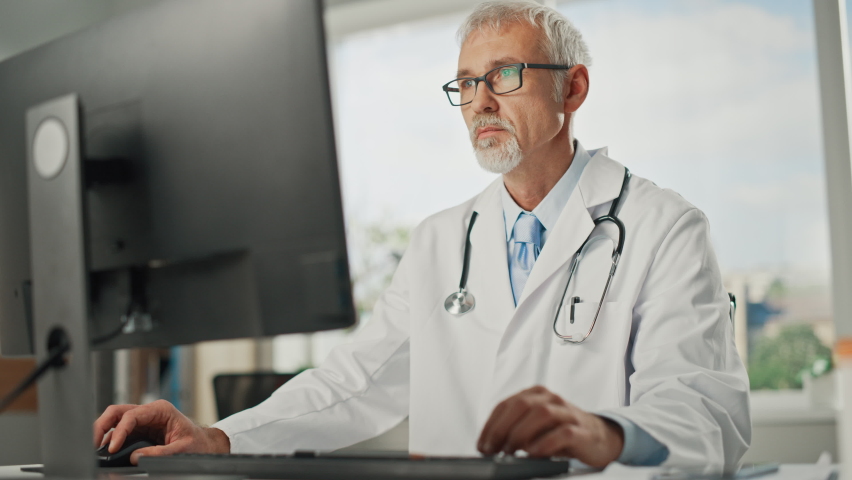 Experienced Middle Aged Male Doctor Wearing White Coat Working on Personal Computer at His Office. Senior Medical Health Care Professional Working with Test Results, Patient Treatment Planning. | Shutterstock HD Video #1065599419