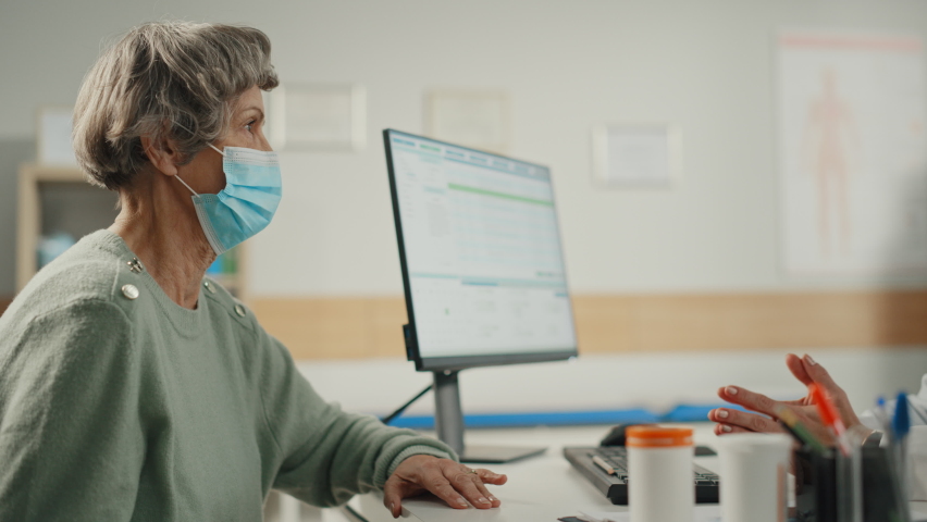 Female Family Doctor is Talking with Senior Old Patient During Consultation in a Health Clinic. Both Wear Face Masks. Physician in Lab Coat Sitting Behind a Computer Desk in Hospital Office. | Shutterstock HD Video #1065599434