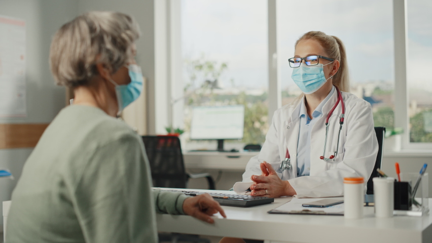 Female Family Doctor is Talking with Senior Old Patient During Consultation in a Health Clinic. Both Wear Face Masks. Physician in Lab Coat Sitting Behind a Computer Desk in Hospital Office. | Shutterstock HD Video #1065599437