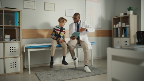 Friendly African American Family Doctor Talking with a Young Boy with Arm Brace and Showing Test Results on Tablet. Happy Medical Care Physician in a Hospital is Reassuring the Boy with Broken Arm.