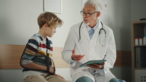 Friendly Middle Aged Family Doctor Talking with a Young Boy with Arm Brace and Showing Test Results on Tablet. Happy Medical Care Physician in a Hospital is Reassuring the Boy with Broken Arm.