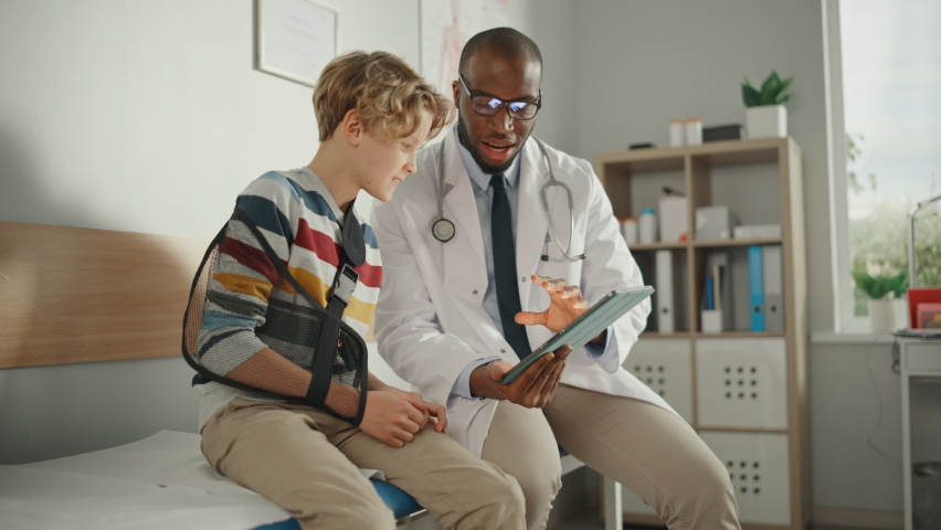 Friendly African American Family Doctor Talking with a Young Boy with Arm Brace and Showing Test Results on Tablet. Happy Medical Care Physician in a Hospital is Reassuring the Boy with Broken Arm. | Shutterstock HD Video #1065599707