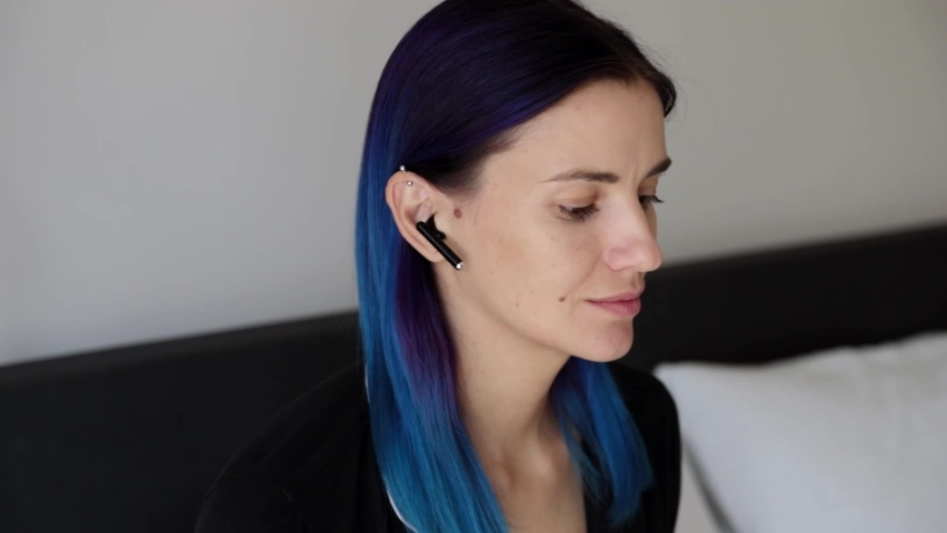 Young woman with blue hair laugh sitting on the bed wearing wireless headphones