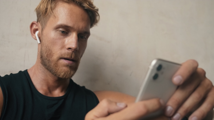 A handsome sportsman with earphones is using his smartphone while sitting near grey wall inside