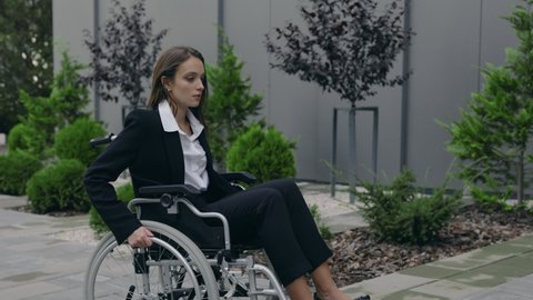 Crop view of disabled female beautiful person in suit pushing herself on wheel chair at street. Invalid woman going to work and stopping near office building stairs. Concept of obstacles, barriers