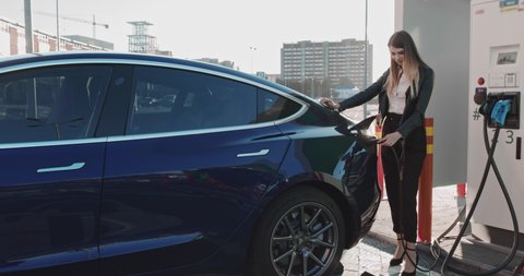 Beautiful Business Woman Relies On A Blue Luxury Electric Car That Is Charging. Electric Car Charging Plug. Luxury Electric Car Is Charging. Girl Looks At The Camera And Smiles. Business Woman Smiling