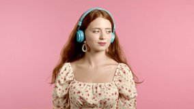Pretty young girl with red hair having fun, smiling, dancing with blue headphones in studio against pink background. Music, dance, radio concept, slow motion