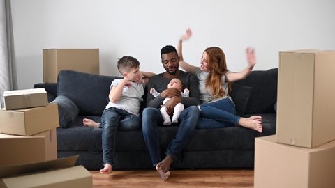 Long-awaited moving in a new house. A multiracial family raising hands up as sign of happy and success. Overjoyed diverse parents, infant and school-age boy sit on the couch among cardboard boxes