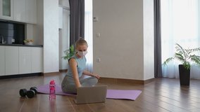 Active young female wearing medical mask while practising yoga at home. Sporty woman in bra and leggings training while watching video on laptop.