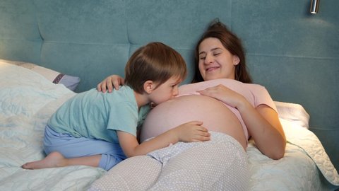 Smiling toddler boy in pajamas talking and hugging unborn baby in big belly of pregnant mother lying in bed at night.
