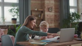 Medium shot of kind woman sitting at desk with small girl teaching drawing pointing at laptop screen while older girl sitting on sofa reading in background