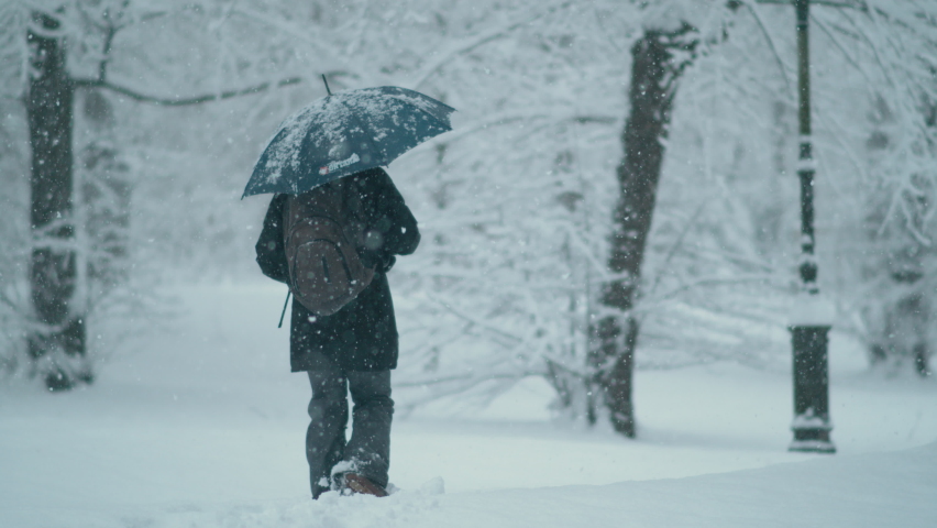 SLOW MOTION, DOF: Unrecognizable woman treads deep powder snow while walking around the wintry park during a blizzard. Female pedestrian walks in the deep snow while exploring the idyllic park.