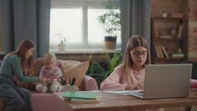Medium shot of young girl sitting at desk studying online via laptop while caring woman playing with kid on sofa in background
