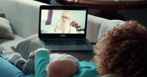 A young mother with a baby is watching online instructions from a female doctor. Baby sleeps on his mother's breast. The doctor advises the young mother in a video call