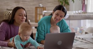 Two sisters with babies follow an online educational program for young mothers, baby exercises and caring for newborn babies. Two little babies lie on their stomachs and look at a laptop. 