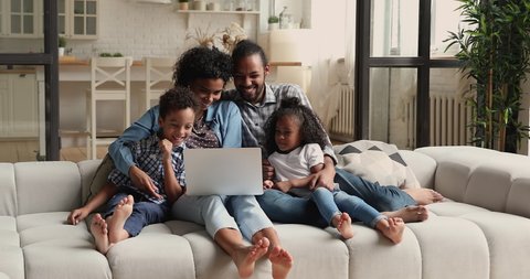 Young African couple little adorable kids relaxing on couch with laptop, family watch funny cartoons, comedy movie laughing feels happy and carefree. Weekend using wireless tech and on-line tv concept