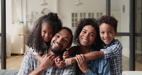 Young african couple with little kids sit on couch, cute children piggyback embrace loving parents smile look at camera. Medical insurance cover or full family, new house and happy homeowners concept