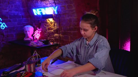 A young beautiful girl makes neon signs in a workshop. A talented artist at work in a multicolored neon studio. The master uses professional equipment, tools and wires.