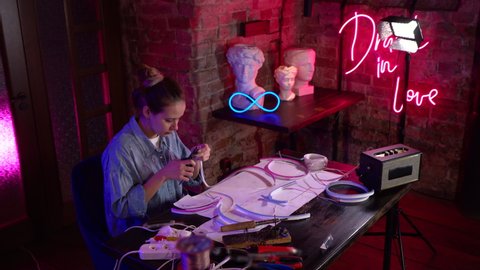A young beautiful girl makes neon signs in a workshop. A talented artist at work in a multicolored neon studio. The master uses professional equipment, tools and wires.