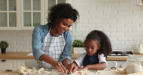 Attractive young African mother and little cute daughter wear aprons cooking together in kitchen, using cookies cutters preparing hand-made biscuits. Hobby, cookery, happy mom teach kid, fun concept