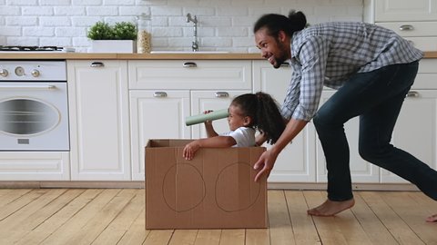 Mixed race African father ride little 5s adorable daughter in carton box, kid girl looking in paper tube binocular enjoy creative game with dad in kitchen. Creativity, handmade plaything, fun concept