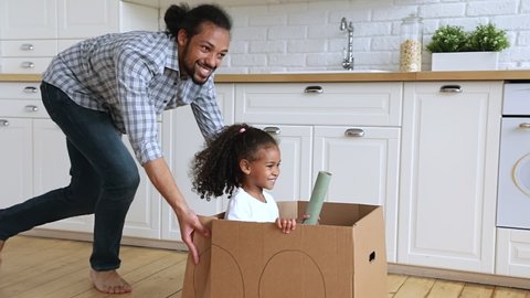 Slow motion African father riding little adorable 4s daughter while she sitting inside of cardboard box, active family play games at modern warm kitchen, celebrate relocation day to new home concept