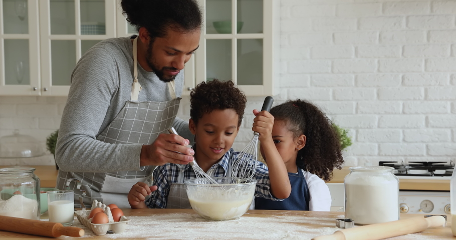 African father and children cooking together in kitchen, kids holding whisks stirring mixture for dough, dad help teaching spend time with lovely son and daughter. Development, hobby, cookery concept Royalty-Free Stock Footage #1065628708