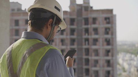 slow motion close up shot of a young male Asian civil engineer wearing hard and safety jacket standing on a top of a building near a construction site using mobile phone to type a text message 