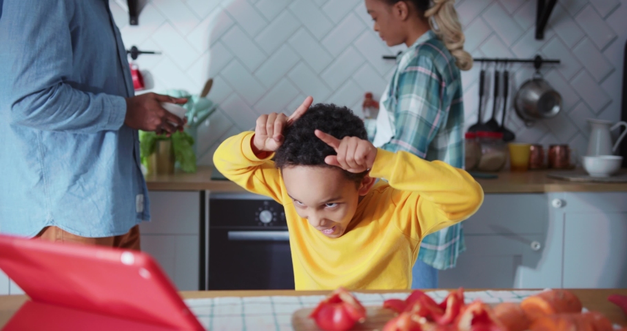 Lovely black young boy acting like cow watching funny cartoons on digital tablet while his parents cooking lunch laughing together. Family in kitchen.