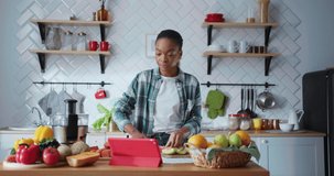Positive african american young woman cooking in her kitche cuttng fruits and talking to mother on online videochat conferece tablet computer.