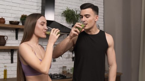 Making green juice at home: Young sporty couple in sportswear drinking smoothie from a glass. Middle shot. Healthy breakfast after a workout in the kitchen 4k