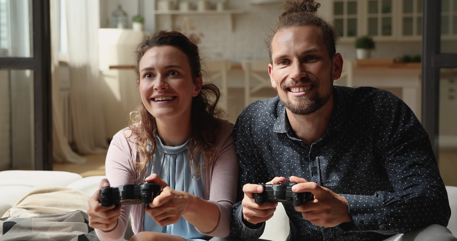Millennial couple holding controllers playing video games sit on couch indoor. Happy family spend weekend enjoy videogame activity feel overjoyed. Have fun, hobby, leisure time at home, gaming concept | Shutterstock HD Video #1065653200
