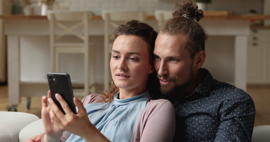 Young millennial couple relaxing on couch with smart phone at home reading sms with great fantastic offer news looking overjoyed celebrate loan bank approve, invitation, good news feeling happiness Royalty-Free Stock Footage #1065653236