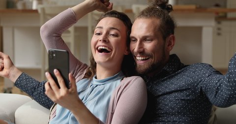 Young millennial couple relaxing on couch with smart phone at home reading sms with great fantastic offer news looking overjoyed celebrate loan bank approve, invitation, good news feeling happiness