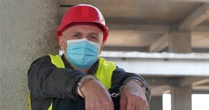 Handcuffed worker in red hard hat and protective face mask sits at construction site and looks at camera. Cinema 4K 60fps video