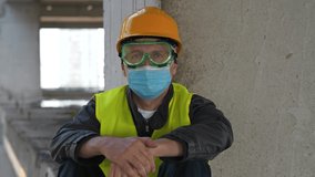 Workman in goggles, hard hat and protective face mask sits at construction site and looks at camera. 4K 60fps panoramic video