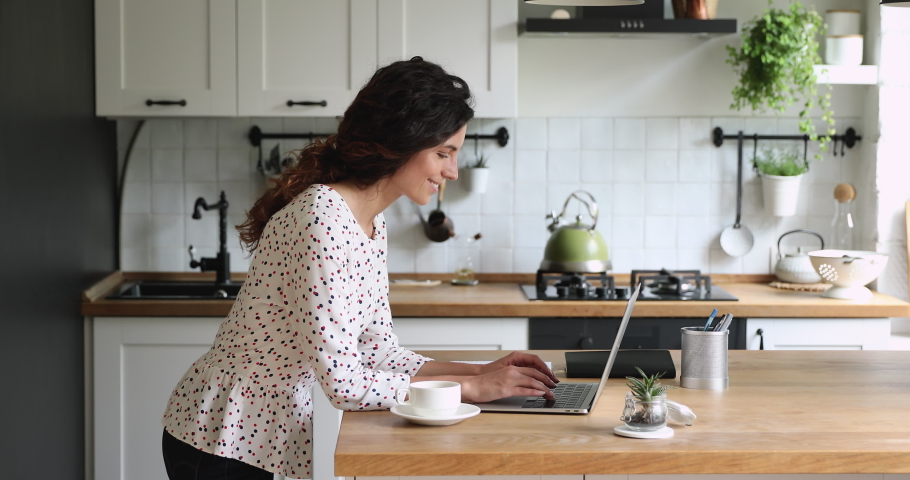 Relaxed young woman drinking hot tea or coffee, web surfing information on computer, communicating with friends in social network, enjoying free weekend peaceful morning time in modern kitchen. | Shutterstock HD Video #1065667591
