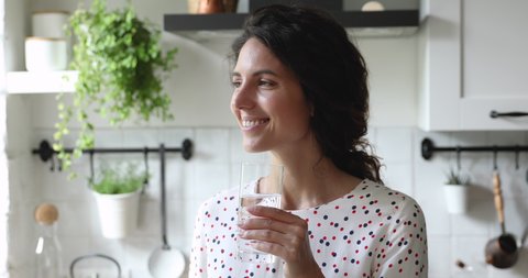 Head shot beautiful young dreamy caucasian woman drinking glass of fresh pure water, enjoying healthy morning habit alone in modern kitchen, starting new energetic day, feeling refreshed and hydrated.